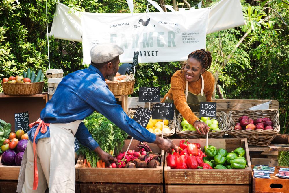 A black woman at a farmers market stand leans over baskets of produce to adjust a sign while a black man places produce in crates. 